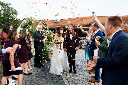 Svatební fotografie novomanželů po obřadu, kteří jdou uličkou mezi hosty. Ti na ně házejí okvětní lístky.